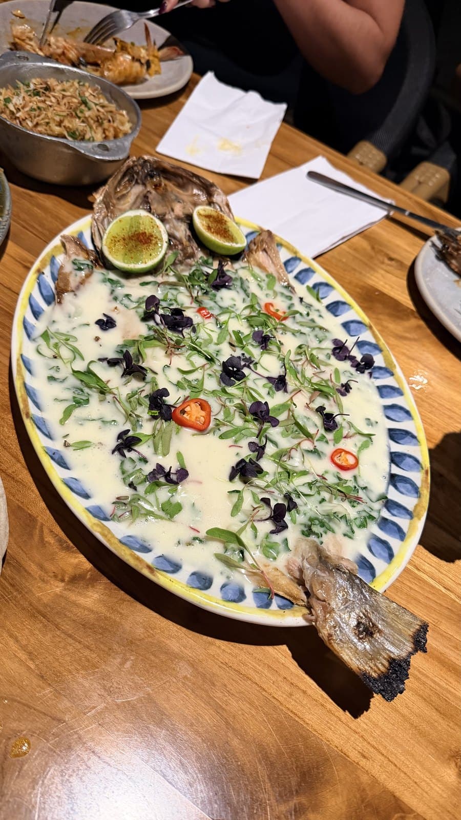 The whole sea bass at Cantina del Tigre on an oval platter, under a pale creamy sauce scattered with micro herbs, sliced chilli and grilled lime